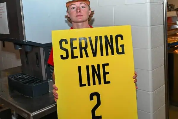 Young volunteer holding a 'Serving Line 2' sign at a charity event.