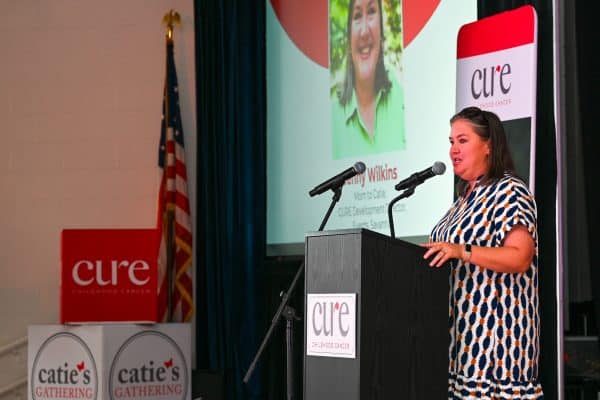 Woman speaking at CURE Childhood Cancer event with presentation screen behind.