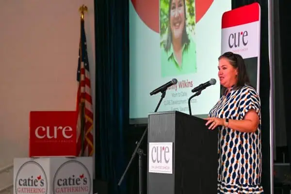 Woman speaking at CURE Childhood Cancer event with presentation screen behind.