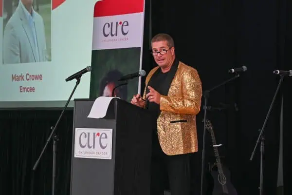 Man speaking at CURE Childhood Cancer event with logo and stage background.