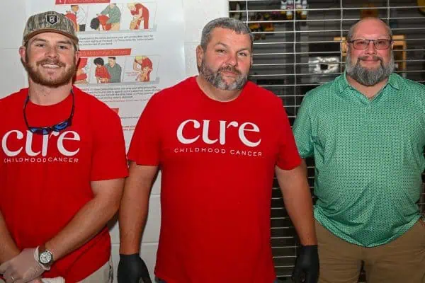 Three men at CURE Childhood Cancer event, smiling and wearing themed shirts.