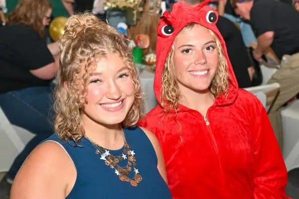 Two smiling women at CURE Childhood Cancer gathering, one in a blue dress, one in a red costume.