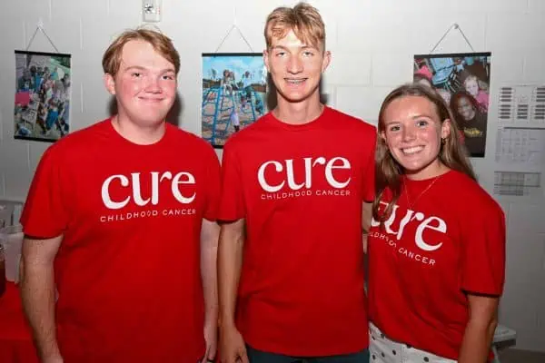 Three young people wearing CURE Childhood Cancer T-shirts at a charity event.