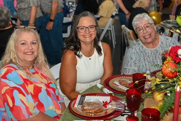 Three women smiling at a charity event table with flowers and food.