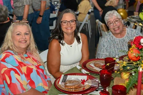 Three women smiling at a charity event table with flowers and food.